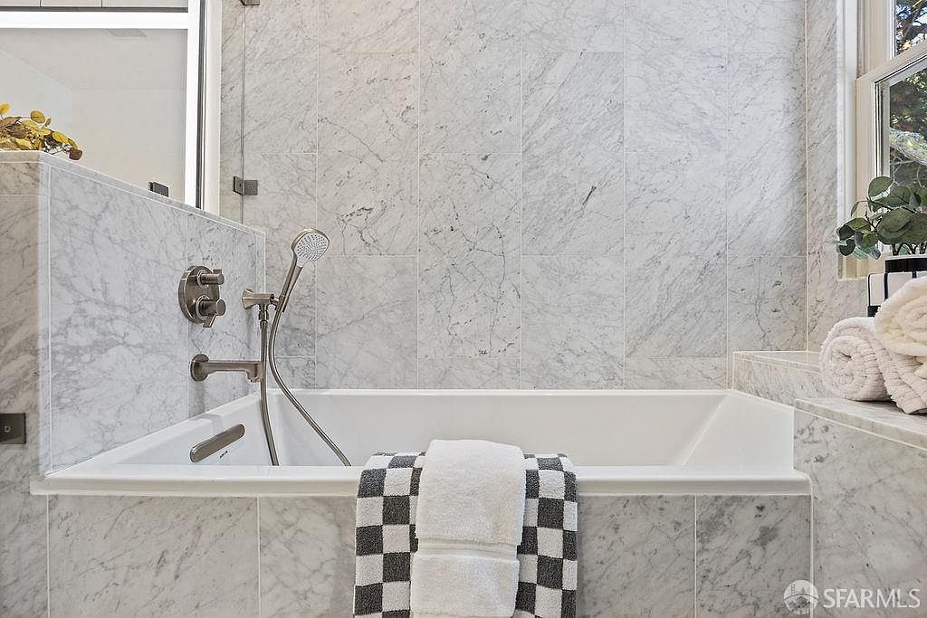This is a primary bathroom featuring a built-in bathtub with marble tile surround. The bathtub has a chrome faucet and shower head, and a white and gray checkered towel is draped over the edge. The walls are covered in marble tiles, and there is a window with a plant and rolled towels on the ledge.