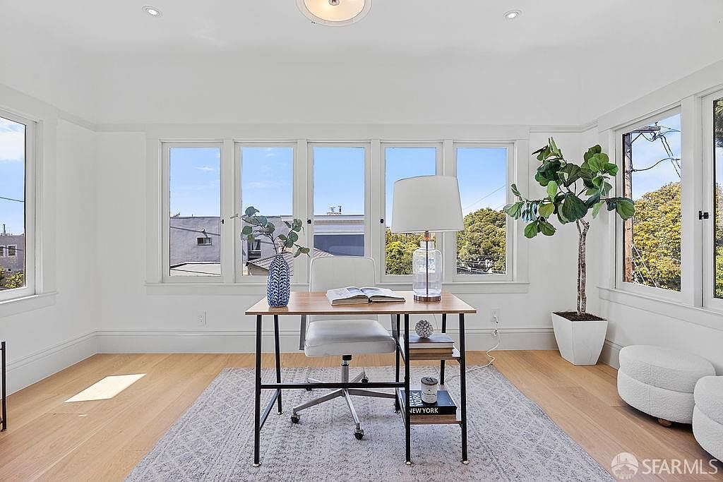 This is an interior shot of a bright and airy office space. The room features a wooden desk with a white chair, a large window providing natural light, and a potted plant adding a touch of greenery. The overall impression is clean, modern, and conducive to productivity.