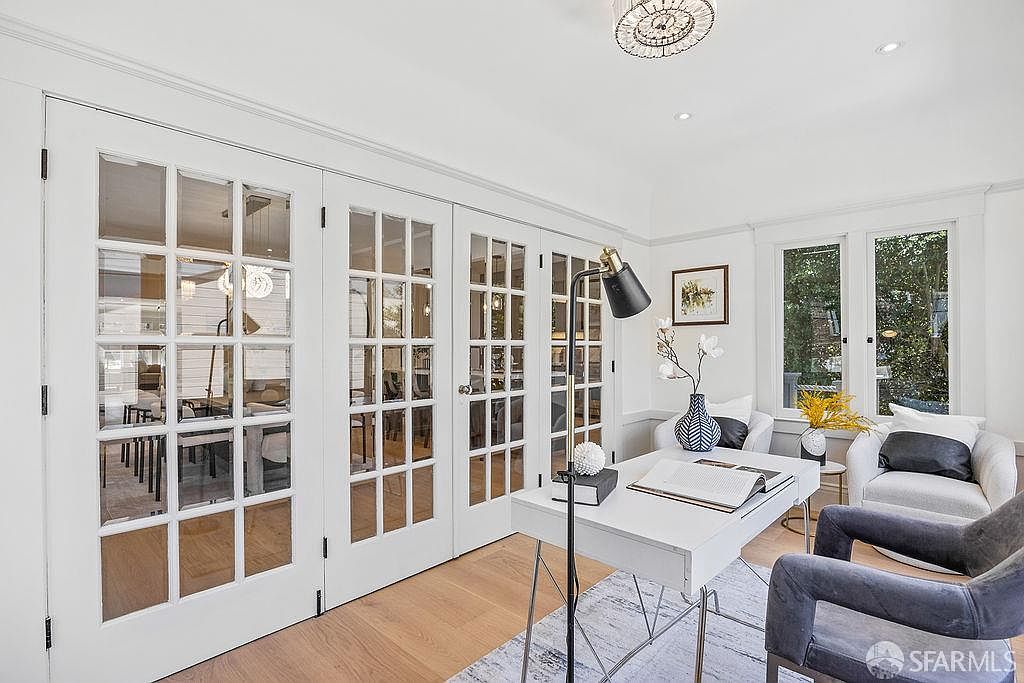 This is an interior shot of a bright and stylish home office. The room features white French doors, a modern white desk with a black lamp, and comfortable armchairs. The hardwood floors and a patterned rug add warmth and texture to the space, creating an inviting and productive atmosphere.