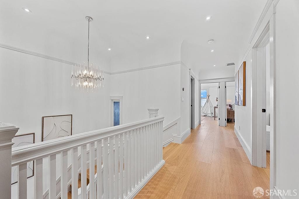 This is an interior shot of a bright hallway and staircase landing. The walls are painted white, and the floors are light wood. A modern chandelier hangs above the staircase, and artwork adorns the walls. The hallway leads to other rooms, suggesting a spacious and well-lit home.