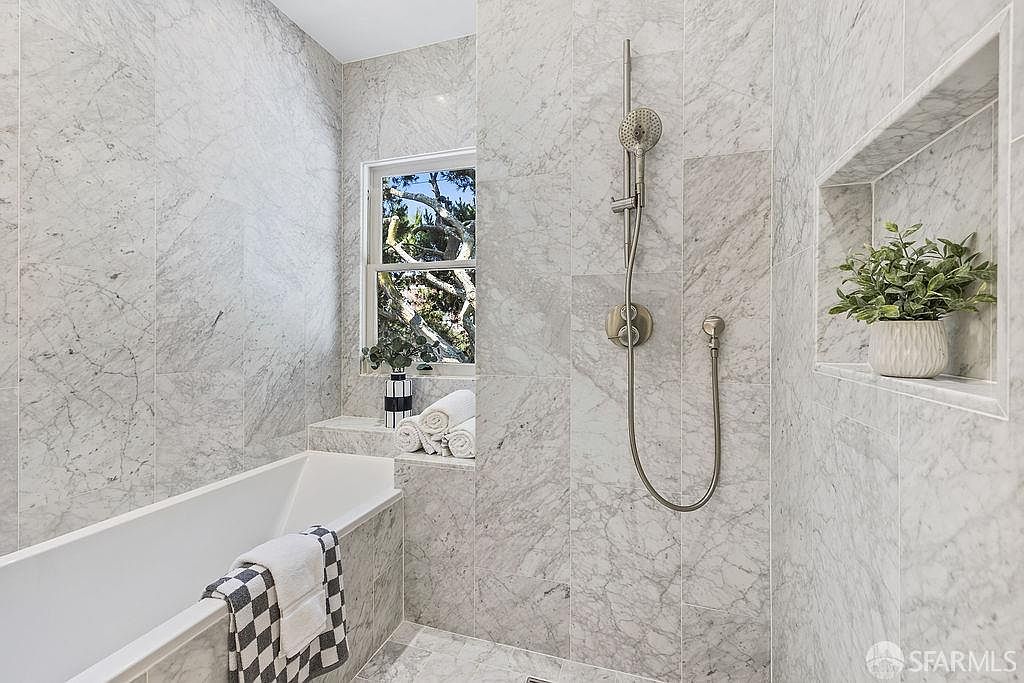 This is a bright and luxurious primary bathroom featuring marble tiling throughout. A freestanding bathtub is positioned next to a window, and a shower area includes a rainfall showerhead and a built-in niche for storage or decor. The overall impression is clean, modern, and spa-like.