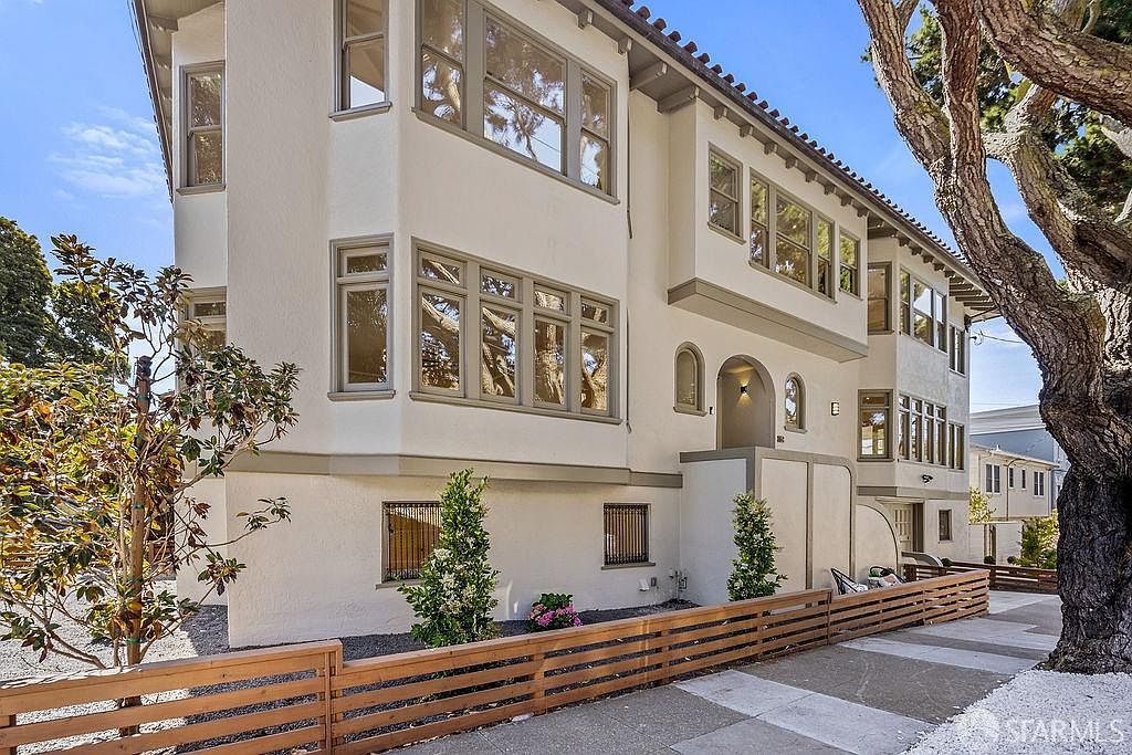 This is a front exterior view of a multi-story building with a white facade and gray trim. The building features numerous windows reflecting the sky and surrounding trees, a tiled roof, and a well-maintained front yard with a wooden fence. The overall impression is of a stately and well-cared-for property.