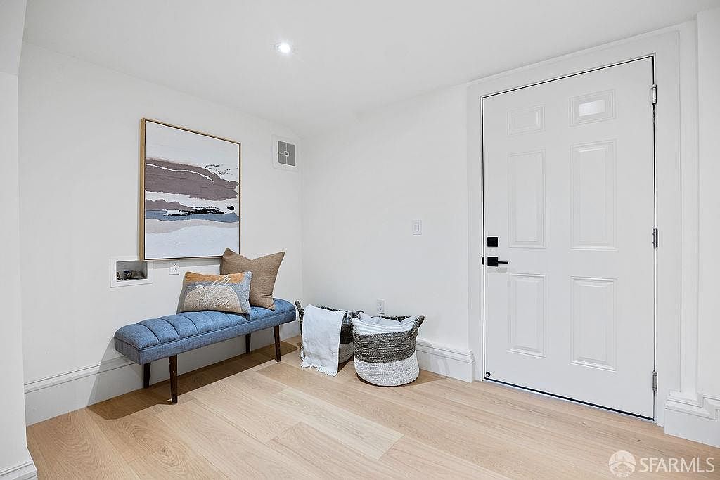 The image showcases a bright and clean hallway featuring light wood flooring and white walls. A blue upholstered bench with decorative pillows sits beneath an abstract art piece, adding a touch of color and comfort. A white door with black hardware is visible on the right, and a woven basket adds a decorative element to the space.