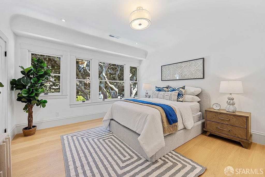 This is a bright and airy guest bedroom featuring a neutral color palette with pops of blue. The room is well-lit by natural light from the windows and a modern ceiling light fixture. A striped rug adds texture to the hardwood floors, and a wooden nightstand with a lamp sits beside the bed, creating a cozy and inviting atmosphere.