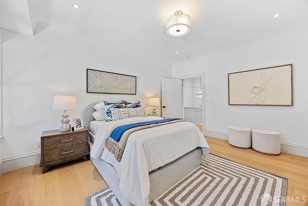 This is a well-lit bedroom featuring a bed with a gray upholstered headboard, adorned with white and blue patterned pillows and a blue throw. A wooden nightstand with a lamp sits beside the bed, and artwork hangs above the bed and on the adjacent wall. The room is finished with a geometric patterned rug and two small upholstered ottomans, creating a cozy and inviting atmosphere.