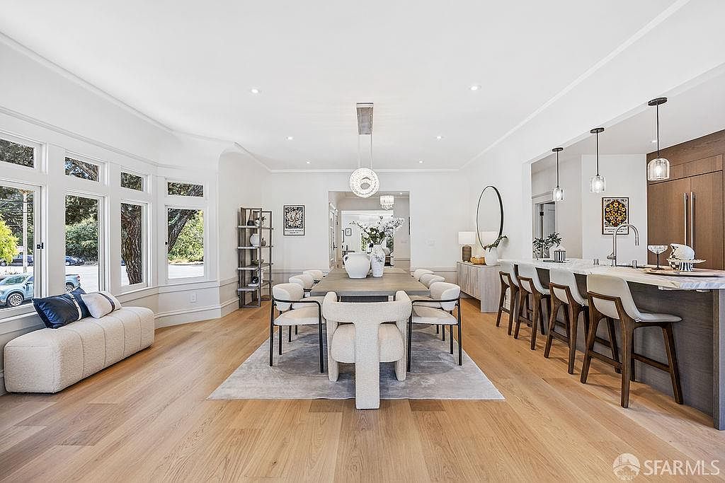This is an interior shot of a dining room that flows into a kitchen area. The dining room features a long table with modern chairs, a light fixture hanging above, and a large window with a bench seat. The kitchen area has a breakfast bar with stools and modern pendant lighting, creating an open and inviting space.