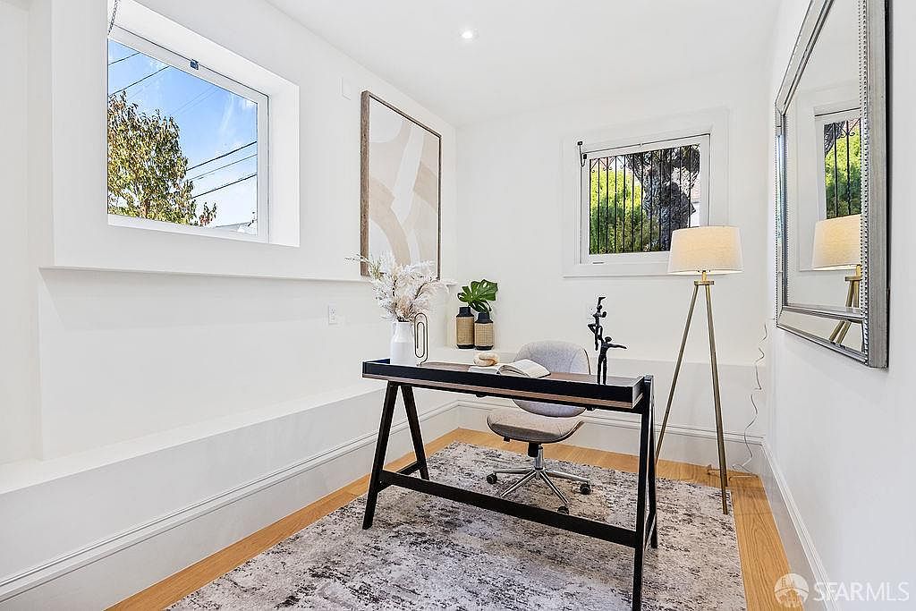 This is an interior shot of a home office featuring a black A-frame desk with decorative items, a gray office chair, and a modern rug. The room has white walls, two windows providing natural light, and a large framed artwork. A floor lamp and a mirror add to the room's ambiance, creating a bright and functional workspace.
