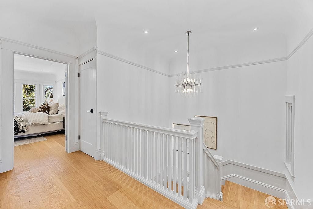 This interior shot showcases a bright hallway with hardwood floors and white walls, leading to a bedroom visible through an open doorway. A white railing lines the staircase, and a modern chandelier hangs from the ceiling, adding a touch of elegance. The space feels clean, airy, and well-maintained, suggesting a move-in ready condition.