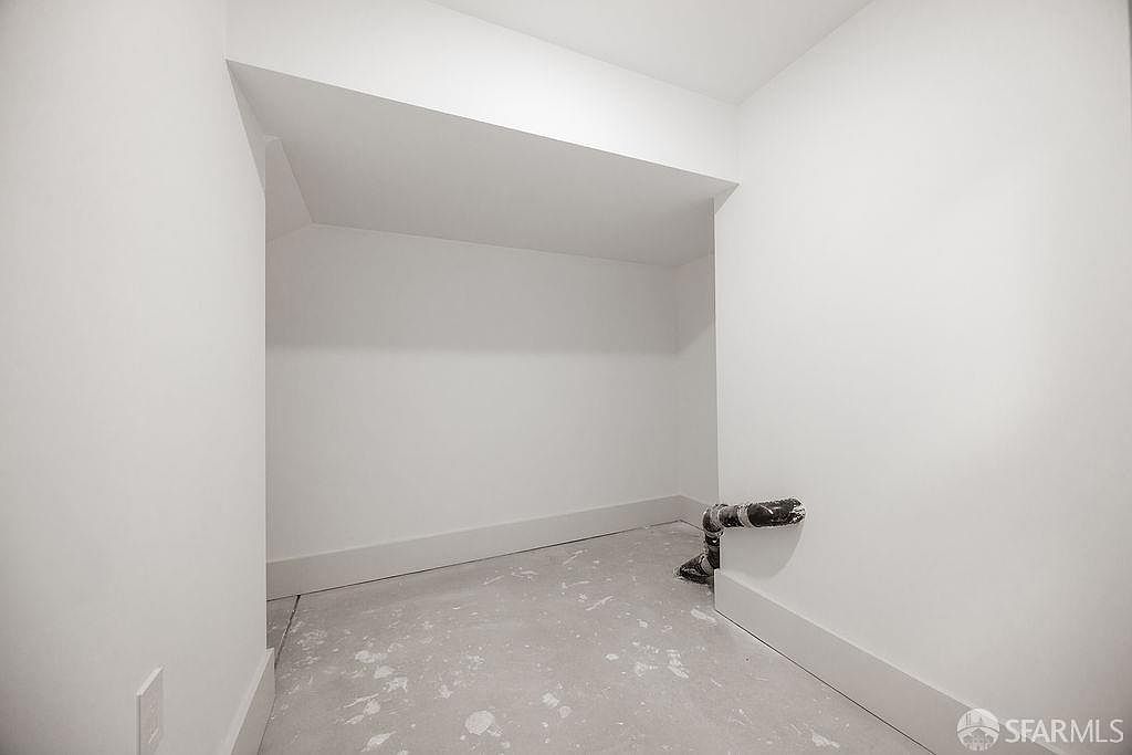 This is a walk-in closet featuring white walls and trim, with a concrete floor that has some paint splatters. A black pipe protrudes from the wall near the floor. The space is bright and appears to be newly constructed or renovated, offering a blank canvas for customization.