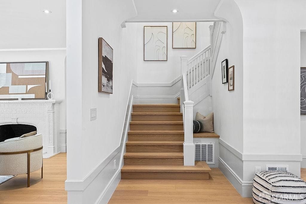 This interior shot showcases a bright and inviting hallway with wooden stairs leading upwards. The walls are painted white, complemented by white trim and baseboards, and adorned with modern artwork. A small seating nook with a pillow is nestled beside the stairs, adding a touch of comfort and style.