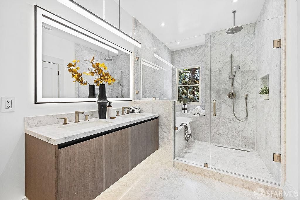 This is a primary bathroom featuring a double vanity with a marble countertop and modern faucets. A large, illuminated mirror hangs above the vanity, and a glass-enclosed shower with marble walls is visible to the right. The bathroom has a clean, luxurious aesthetic with a neutral color palette.