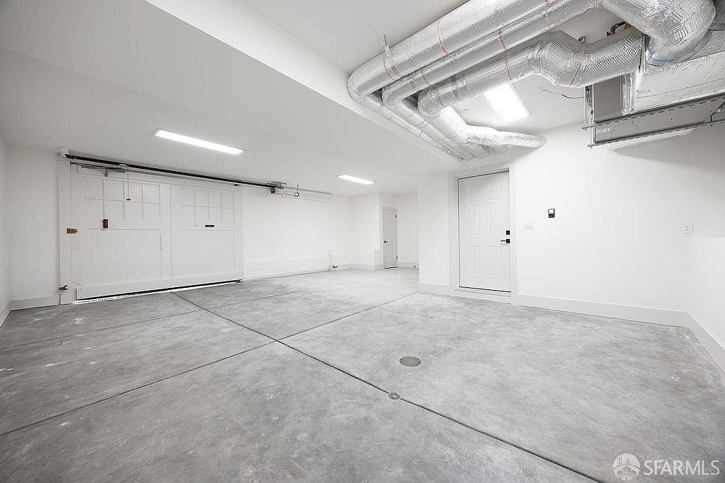 This is a spacious and well-lit garage interior. The walls and ceiling are painted white, creating a clean and bright atmosphere. The floor is concrete with defined sections, and there's a large garage door on one side, along with a standard door leading to the interior of the house. Exposed ductwork adds an industrial touch.
