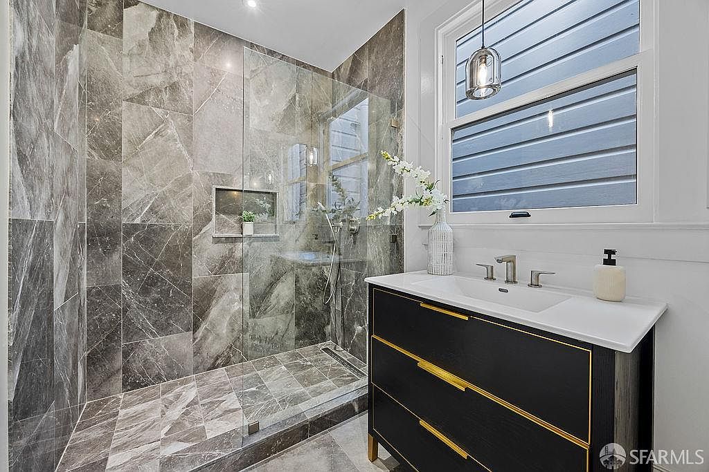 This is a stylish bathroom featuring a walk-in shower with gray marble-like tiling and a glass enclosure. The vanity is a dark wood with gold accents and a white countertop, complemented by modern fixtures. A window provides natural light, enhancing the clean and contemporary aesthetic of the space.
