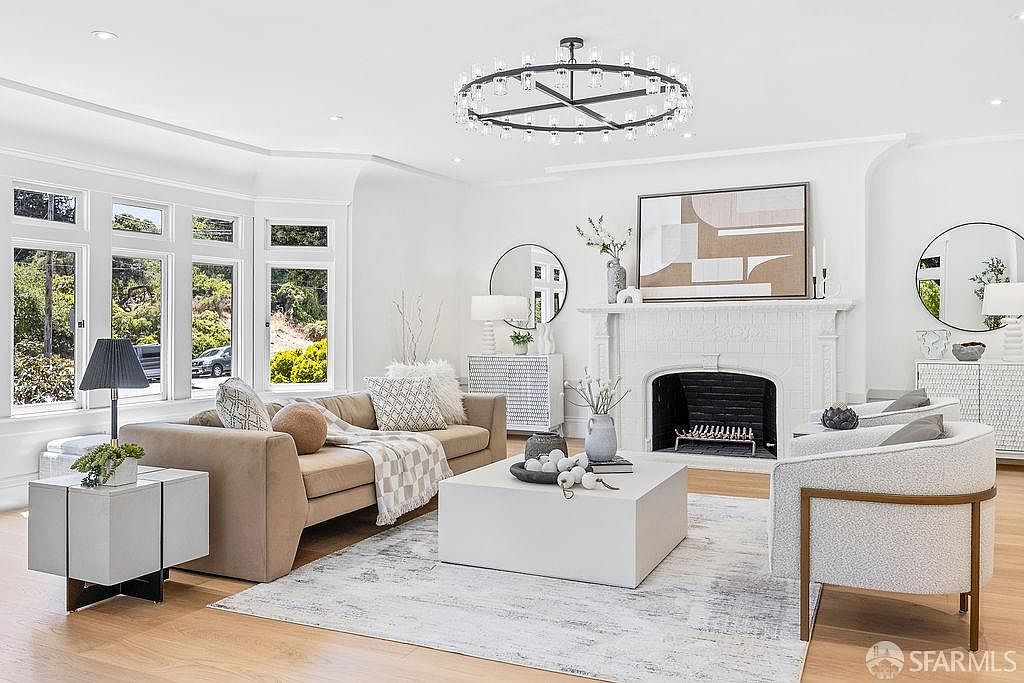 This is a bright and airy living room featuring a neutral color palette. The room is furnished with a beige sofa, a white armchair, and a large white coffee table, all set on a light-colored rug. A fireplace with a white mantel and a large chandelier add to the room's elegance, while large windows provide ample natural light.