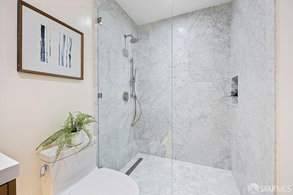 This is a bathroom featuring a modern shower with marble tiling and a glass enclosure. A toilet with a plant on top is visible in the foreground, and a framed artwork hangs on the wall. The overall impression is clean, bright, and stylish.