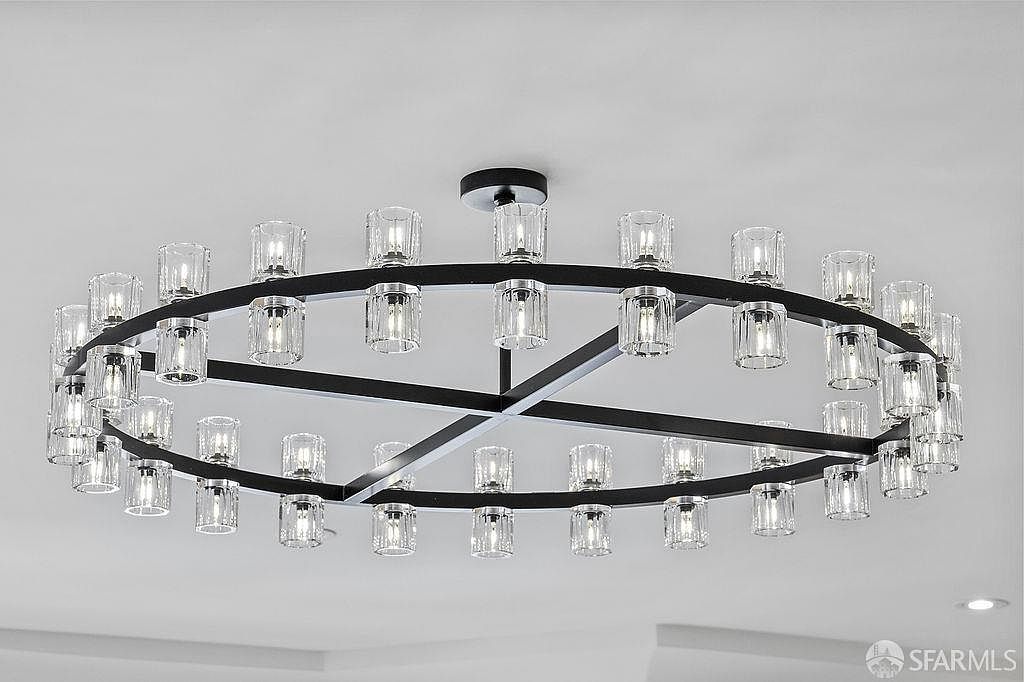The image showcases a modern, circular chandelier with a black metal frame and numerous clear glass shades, each housing a light bulb. The fixture is mounted on a white ceiling, creating a clean and contemporary aesthetic. The perspective is from below, emphasizing the chandelier's design and size.