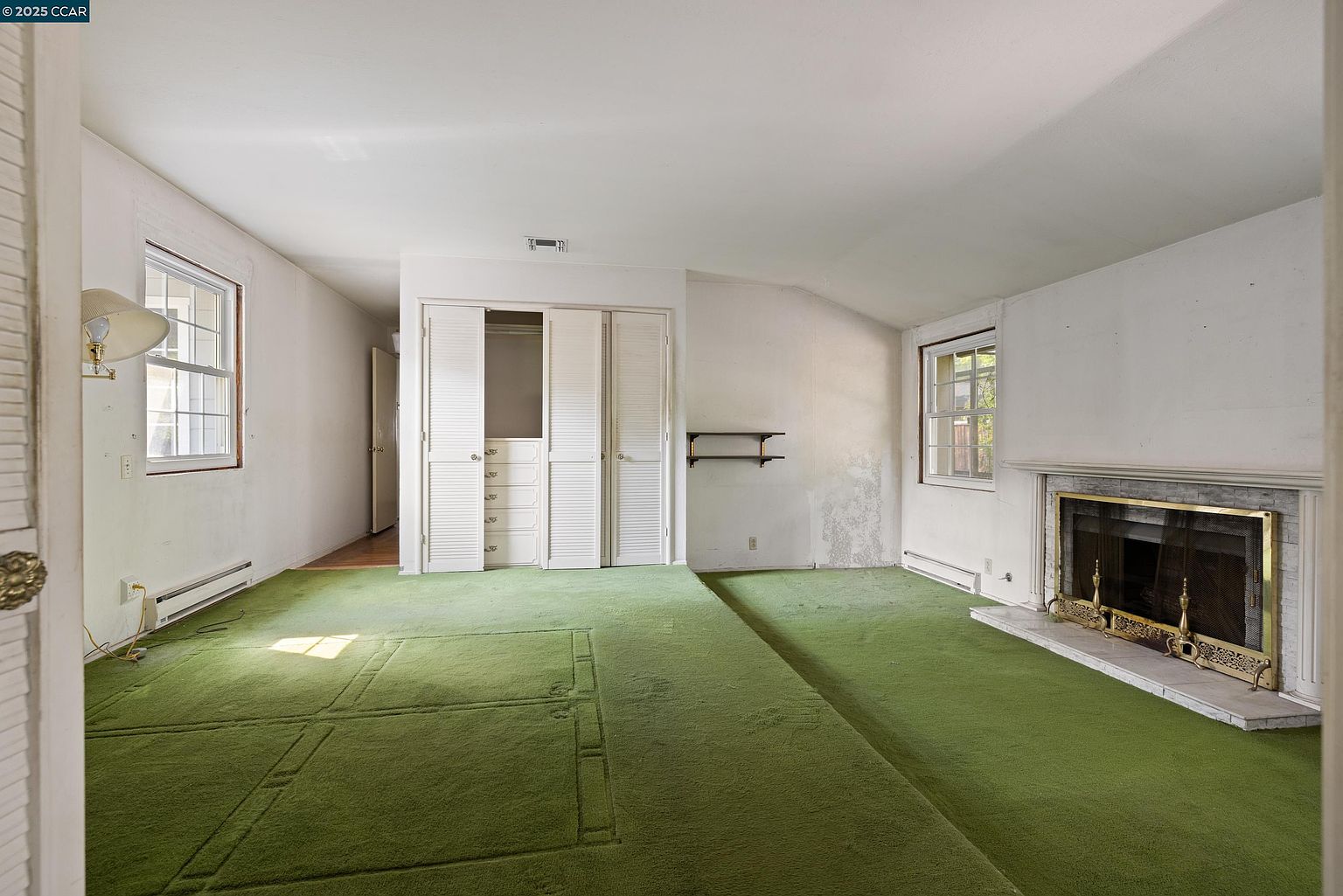This interior shot showcases a bedroom with green carpeting and white walls. The room features a fireplace, a window, and a closet with bi-fold doors. The overall impression is dated but spacious, offering potential for renovation and personalization.