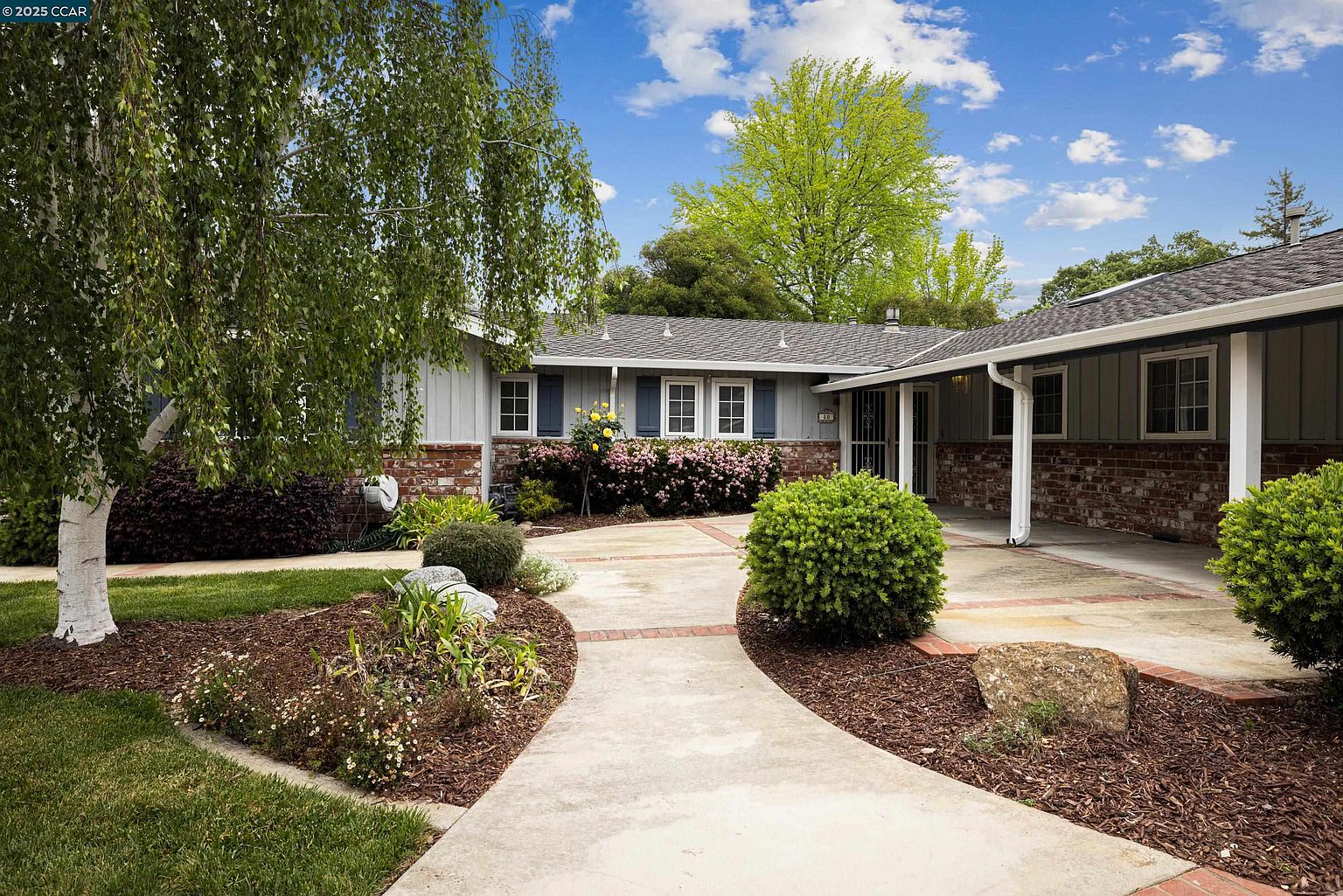 This is a front view of a single-story home with a well-maintained yard and garden. The house features a combination of light gray siding and brick accents, complemented by dark shutters and a gray roof. A curved concrete walkway leads to the front entrance, flanked by landscaped beds with mulch, rocks, and various plants, creating an inviting curb appeal.