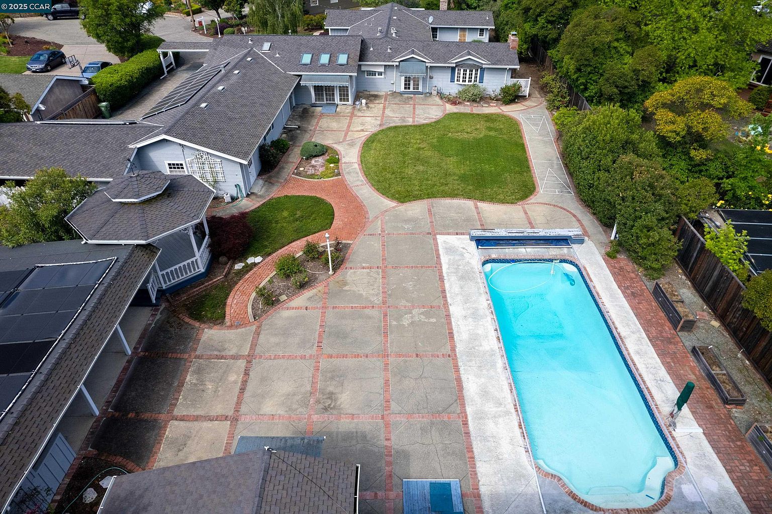 This aerial view showcases a sprawling single-family home with a large backyard featuring a swimming pool, a well-manicured lawn, and a spacious patio area. The house has a light blue exterior with a gray roof, and solar panels are visible on one section of the roof. The perspective is from above, providing a comprehensive view of the property's layout and outdoor amenities.