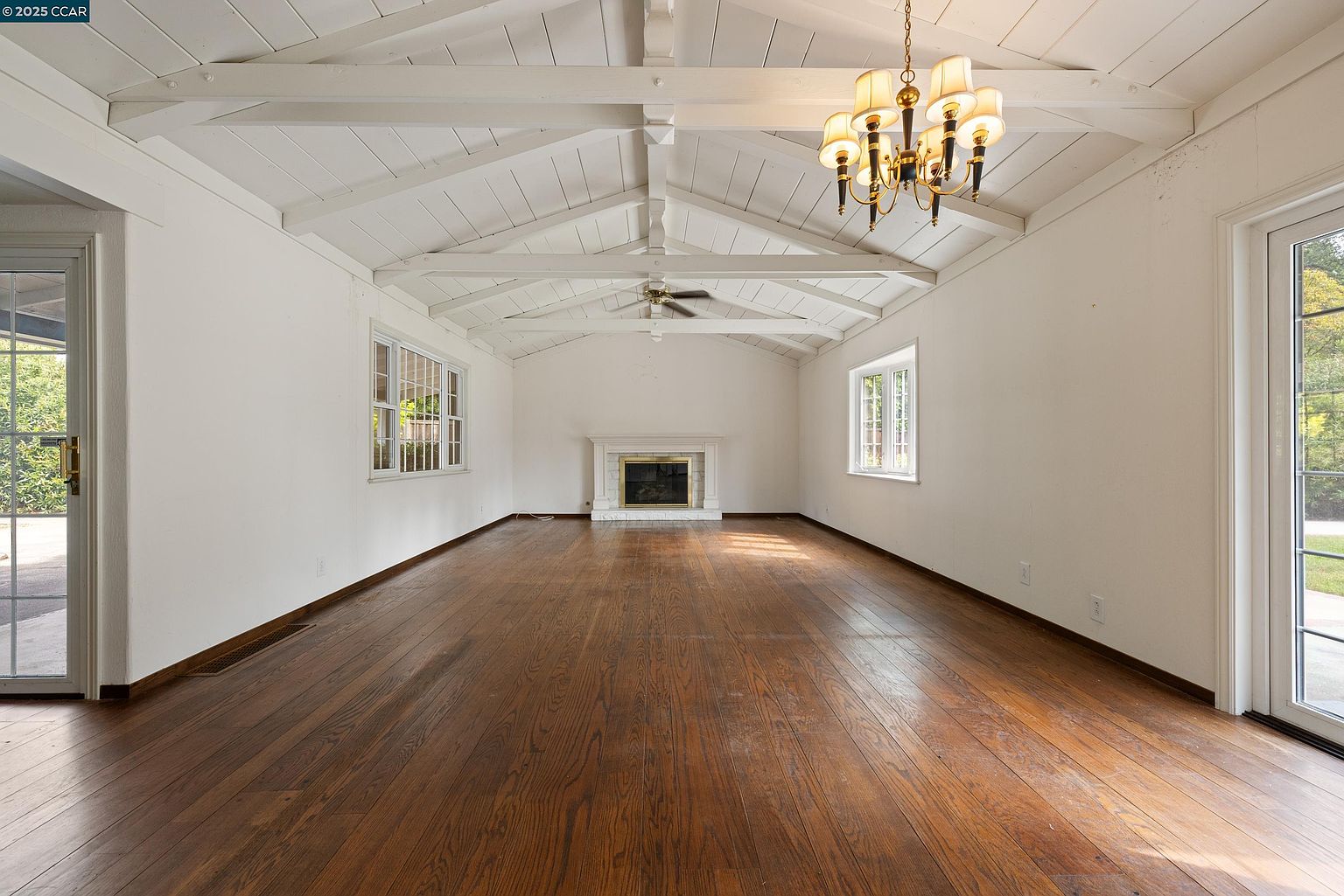 This is an interior shot of a spacious living room featuring hardwood floors, white walls, and a vaulted ceiling with exposed beams. A fireplace is centered on one wall, and natural light streams in through windows and glass doors. A chandelier hangs from the ceiling, adding a touch of elegance.