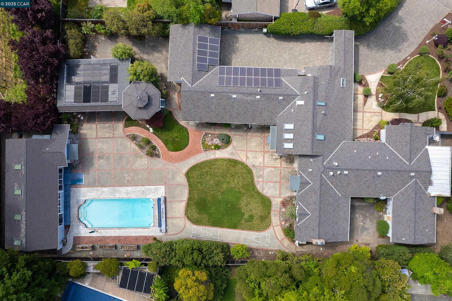 This aerial view showcases a sprawling estate with a large swimming pool, extensive patio area, and well-manicured lawn. The house features a complex roofline with solar panels and multiple wings, suggesting a spacious and luxurious interior. Mature trees and landscaping surround the property, providing privacy and enhancing its curb appeal.