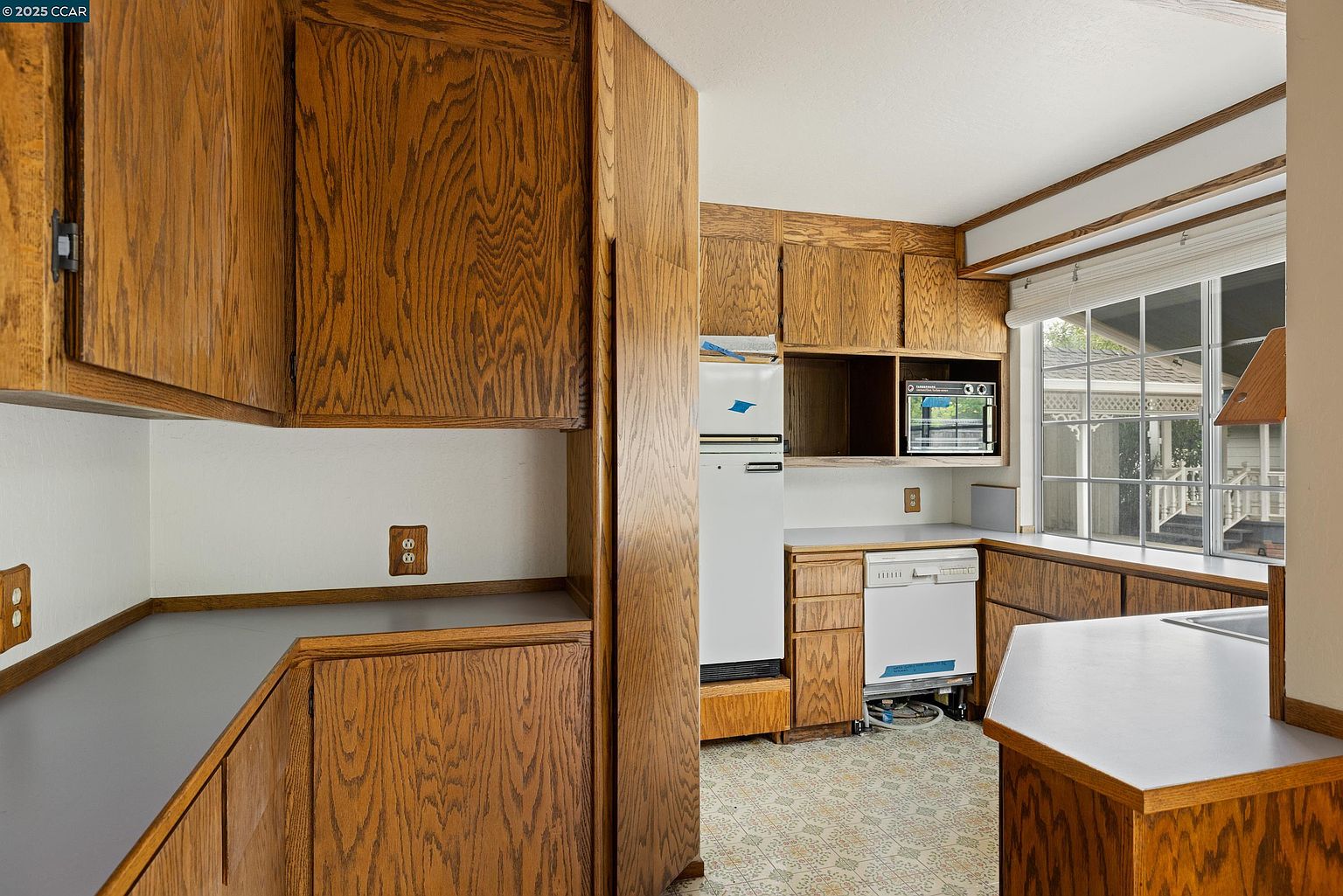 This is a kitchen with wood cabinets and a white countertop. The appliances are white, including a refrigerator and dishwasher. A window provides natural light, and the floor has a patterned design.