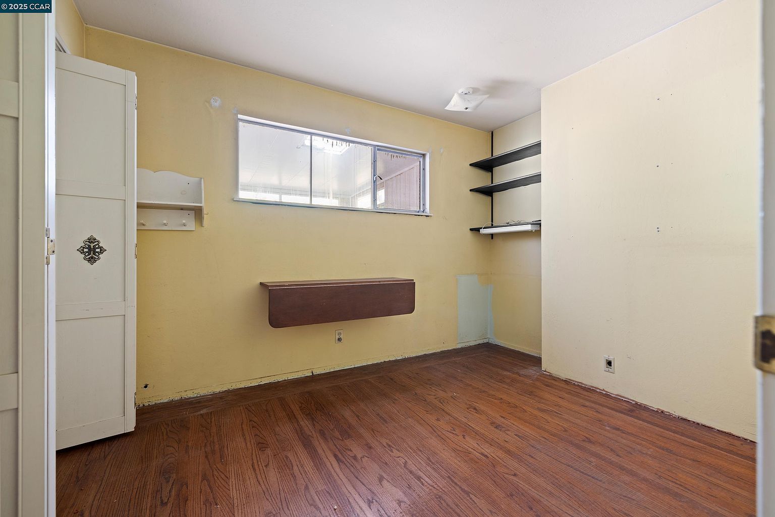 The image showcases a bedroom with hardwood floors and yellow walls. A built-in wardrobe is on the left, while a window and a drop-leaf table are on the back wall. The room also features built-in shelves on the right wall, creating a functional and cozy space.
