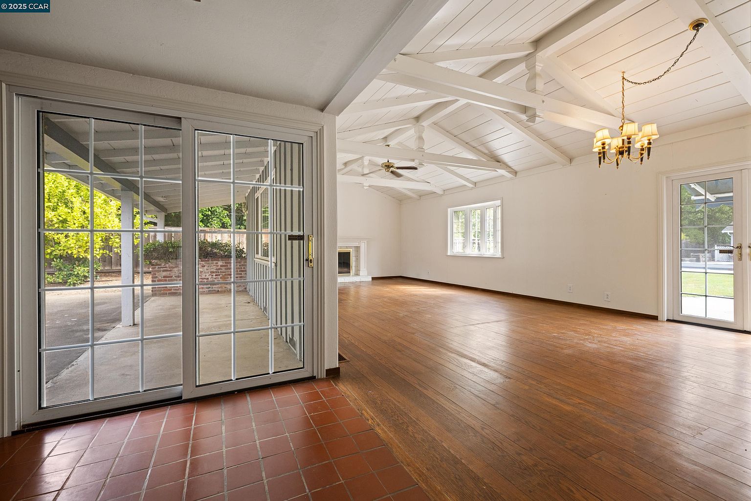 This interior shot showcases a spacious living room with hardwood floors and a high, beamed ceiling. Natural light floods the room through large sliding glass doors and a window, highlighting the room's open layout and potential for various furniture arrangements. A chandelier adds a touch of elegance to the bright and airy space.