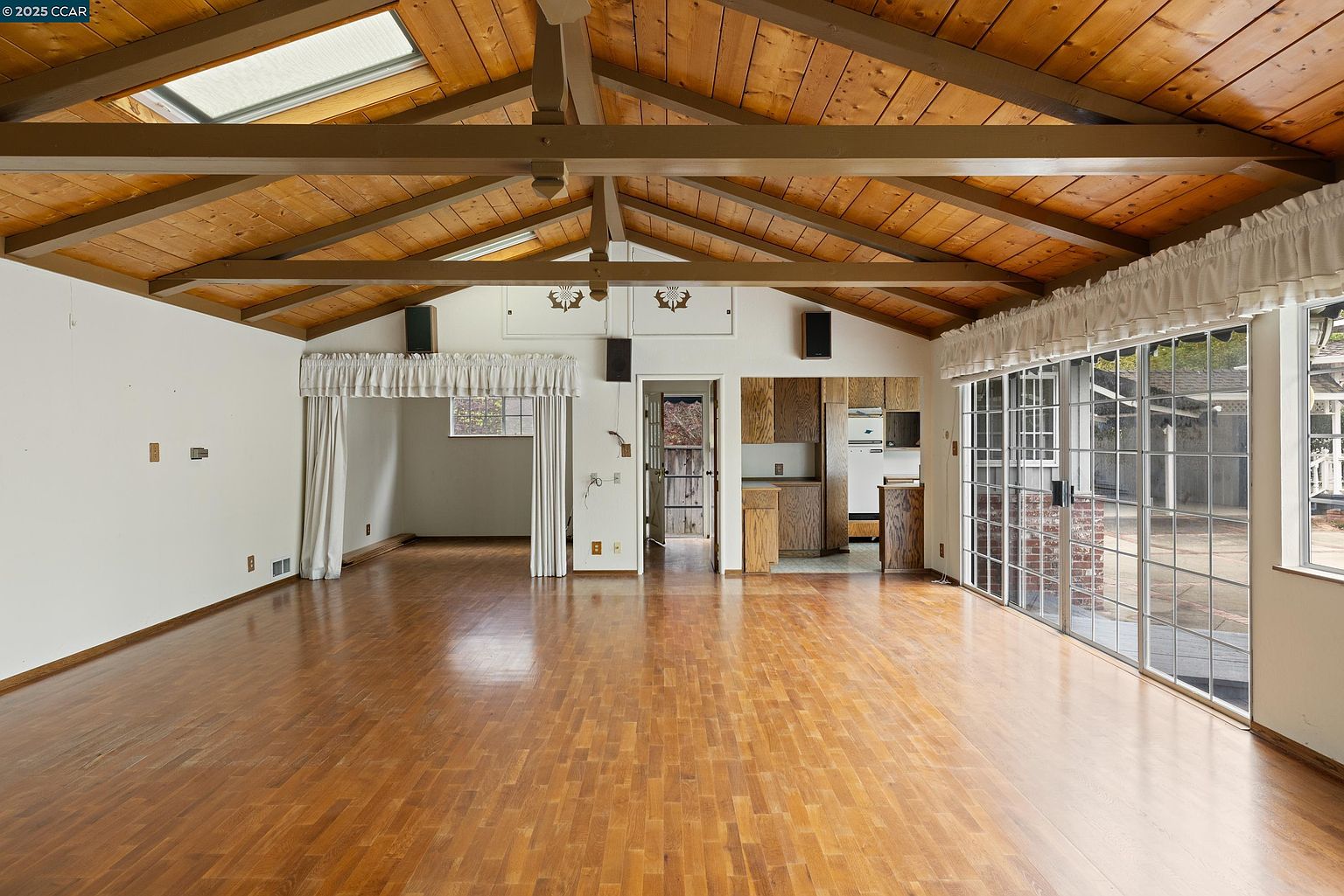 This interior shot showcases a spacious living room with hardwood floors and a high, beamed ceiling featuring skylights. The room opens to an outdoor area through large sliding glass doors, providing ample natural light. The space also includes a partial view of the kitchen area and an adjacent room, suggesting an open floor plan.