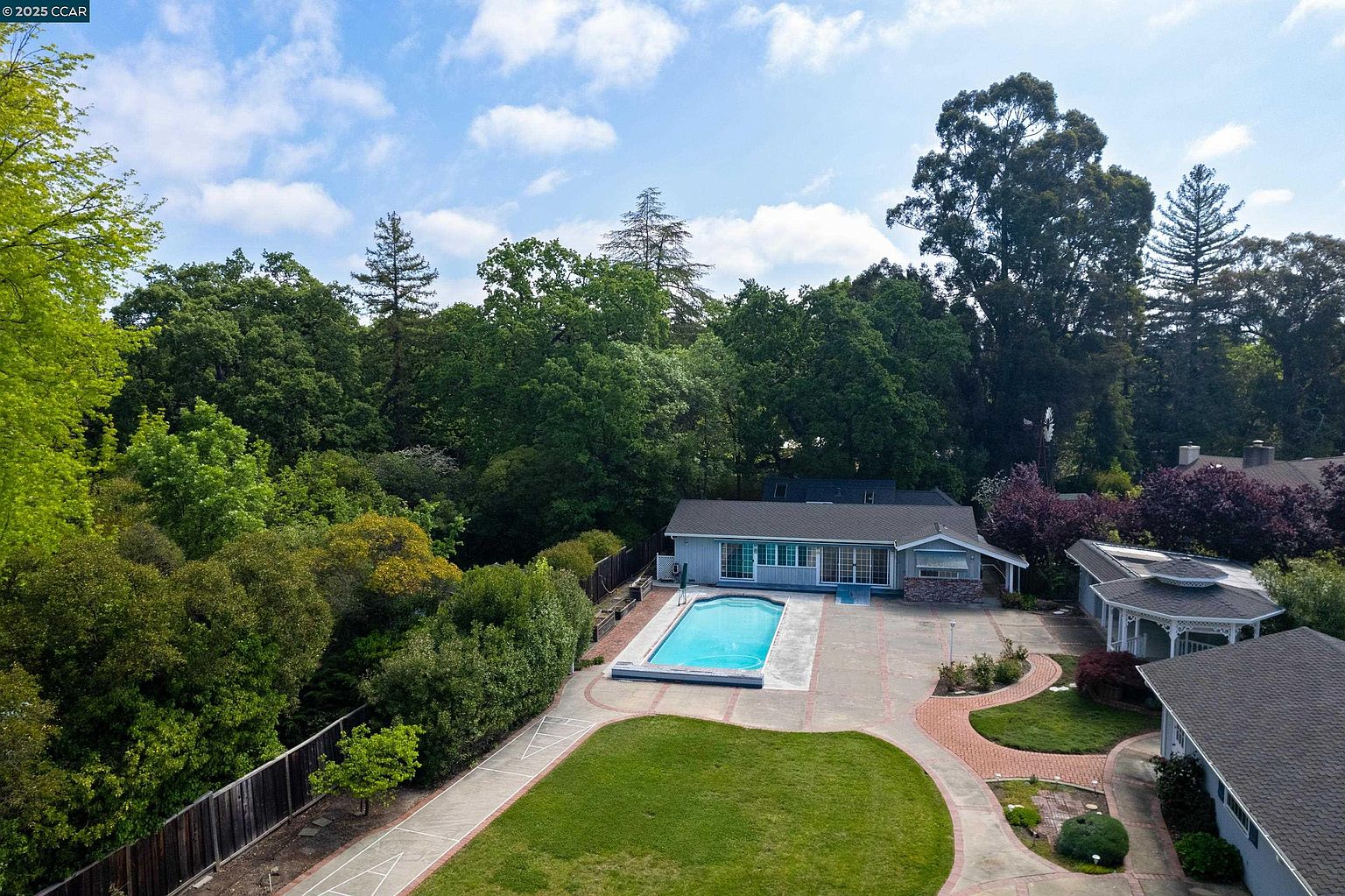 This aerial view showcases a well-maintained property featuring a light blue single-story home with a swimming pool and a spacious brick patio. Lush green lawns and mature trees surround the house, providing privacy and a serene atmosphere. The image highlights the property's outdoor living spaces and landscaping.