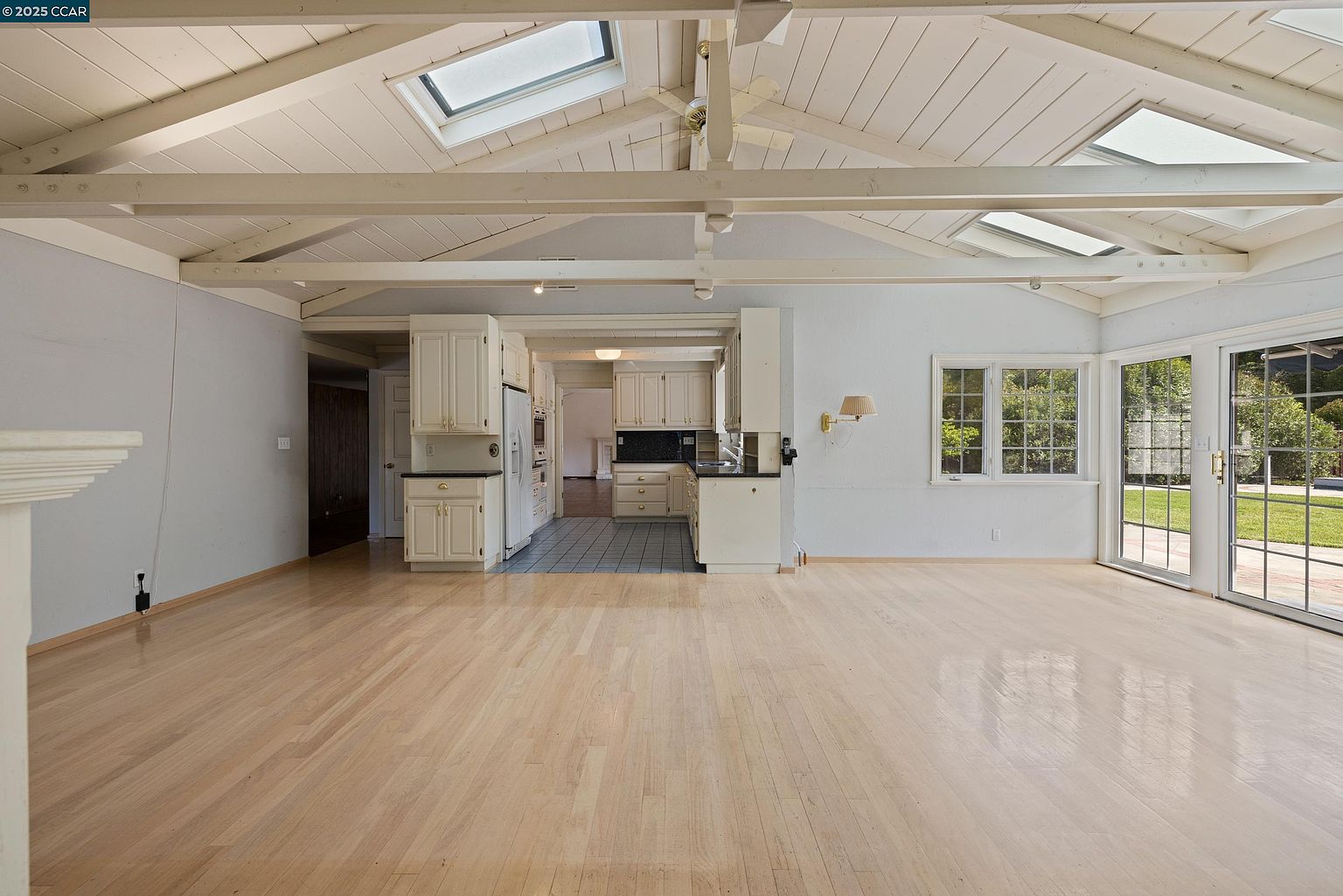 This interior shot showcases a spacious living room with light hardwood floors and a high, vaulted ceiling featuring exposed beams and skylights. The room connects seamlessly to the kitchen area, with white cabinetry visible in the background. Large windows and a sliding glass door provide ample natural light and a view of the exterior.