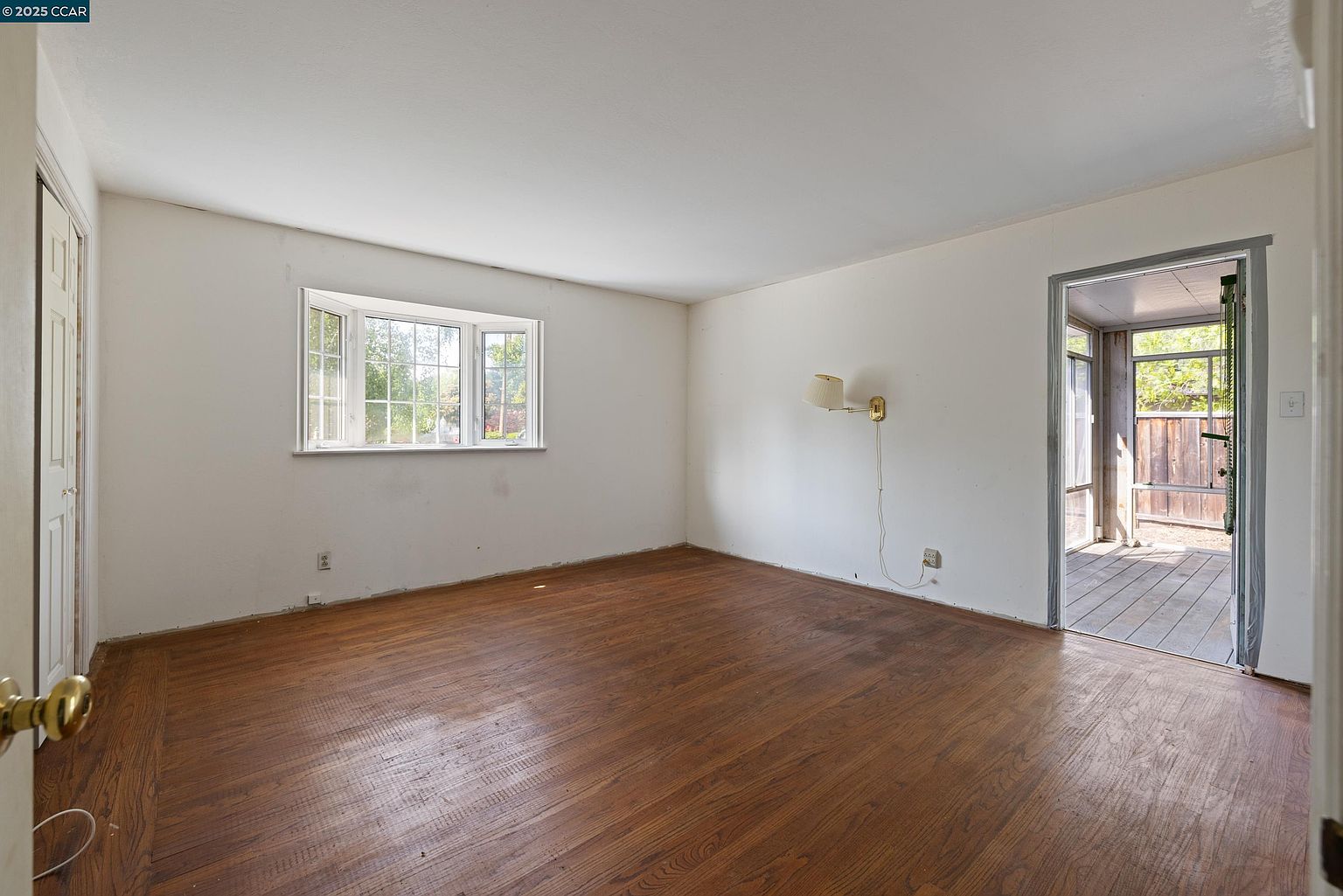 The image showcases an interior bedroom featuring hardwood flooring and white walls. A bay window provides natural light, and a doorway leads to an outdoor patio or deck area. The room appears spacious and ready for personalization, with a simple wall-mounted light fixture adding a touch of character.