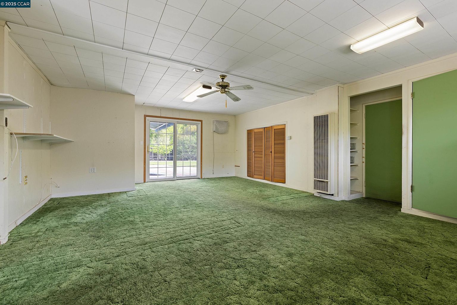 This interior shot showcases a spacious living room with green carpeting and neutral-toned walls. A sliding glass door provides natural light and access to the outside, while a ceiling fan adds to the room's functionality. The room also features built-in storage and a wall-mounted heater, creating a comfortable and practical living space.