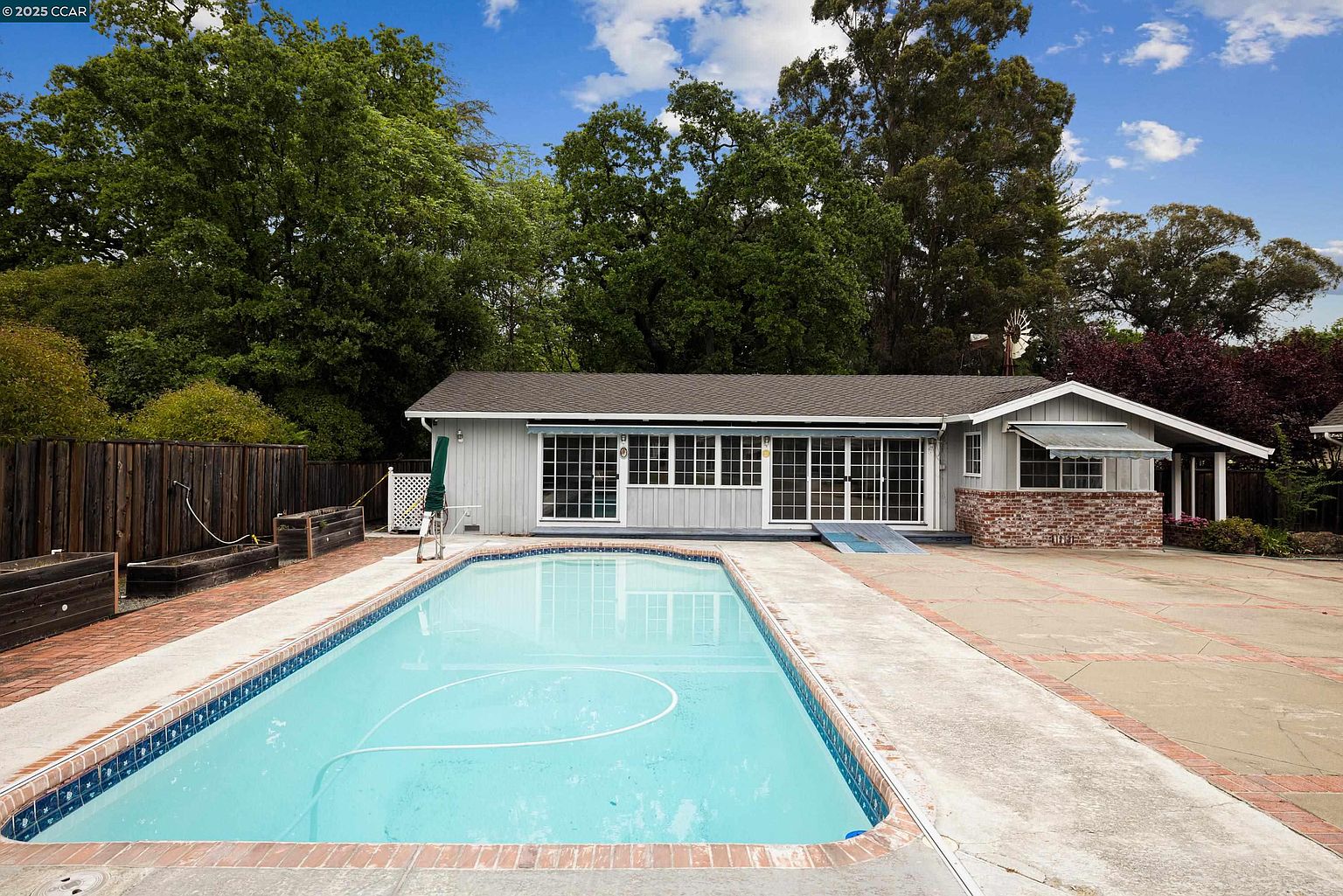 This image showcases a rectangular swimming pool with clear blue water, surrounded by a concrete patio with brick edging. The pool is positioned in front of a single-story house with a sunroom featuring multiple windows and sliding glass doors. Lush green trees provide a backdrop, creating a private and serene outdoor living space.