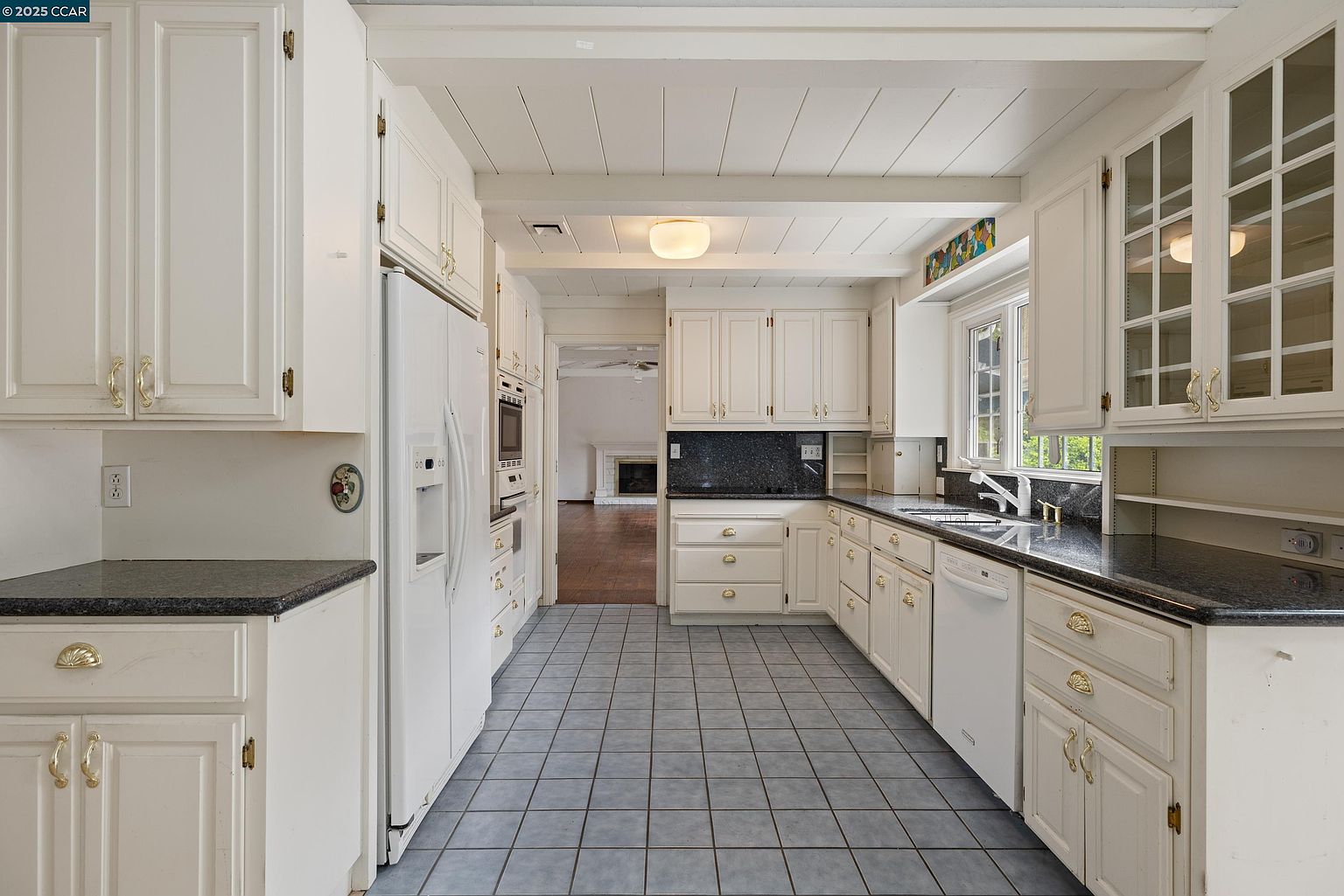 This is a well-lit kitchen featuring white cabinetry with gold hardware, dark countertops, and blue-gray tiled flooring. The kitchen includes a refrigerator, oven, dishwasher, and a window above the sink. The perspective is from the entrance of the kitchen, showcasing the layout and appliances.