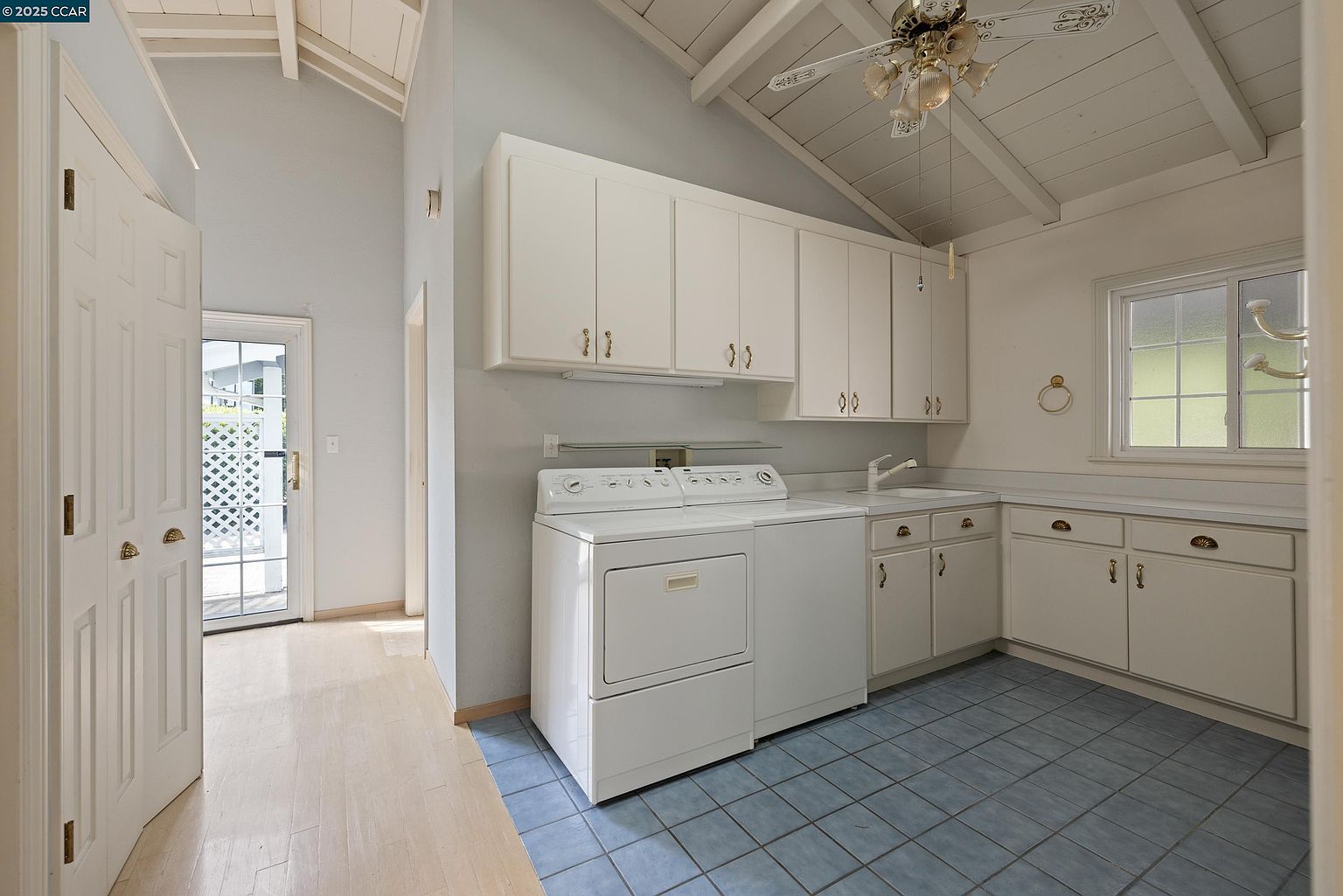 This is an interior shot of a laundry room featuring white cabinetry and appliances. The room has a washer and dryer set, a sink, and ample counter space. The blue tile flooring adds a touch of color to the otherwise neutral palette, and a window provides natural light.