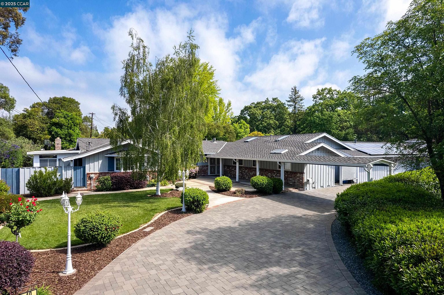 This is a front exterior view of a well-maintained single-story home. The property features a brick and siding facade, a gray roof, and a paved driveway leading to a covered carport. The landscaping includes mature trees, manicured bushes, and a green lawn, enhancing the curb appeal of the residence.