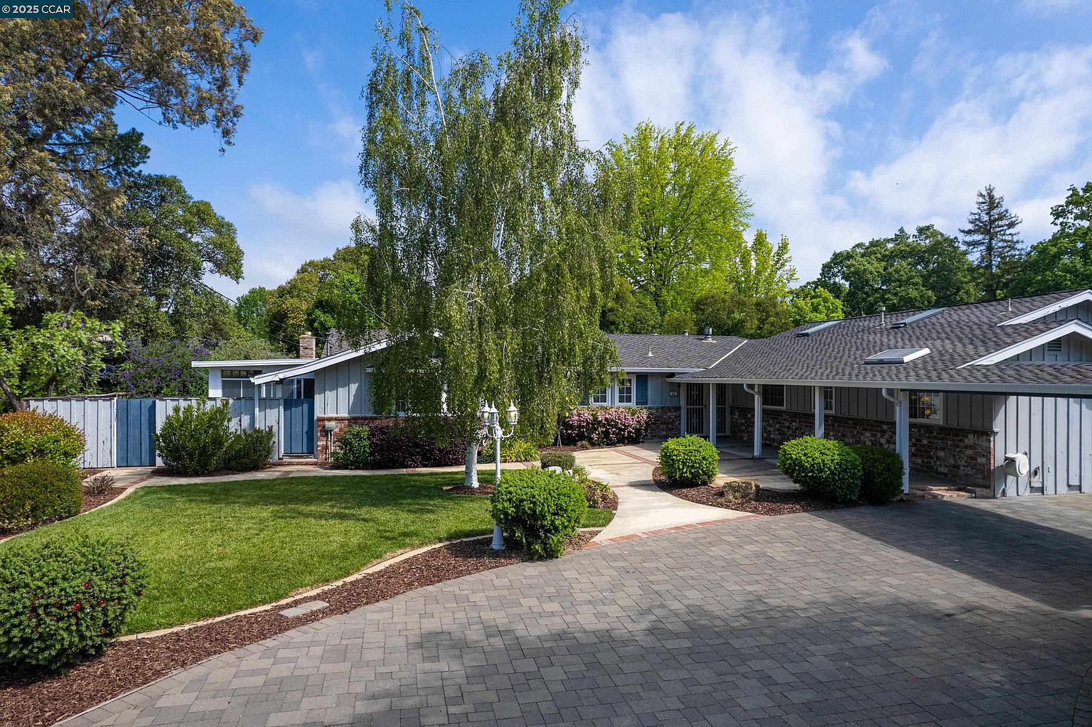 This is a front view of a single-story home with a well-manicured lawn and mature landscaping. The house features a combination of siding and brick accents, a covered entryway, and a paved driveway leading to a garage. A large tree in the front yard adds to the property's curb appeal.