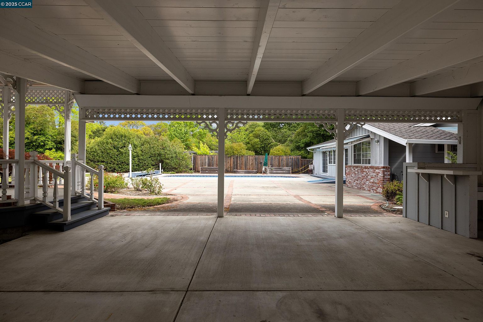 This image showcases a covered patio area with a view of a backyard featuring a pool. The patio has a concrete floor and is supported by decorative white columns and latticework. The backyard includes a pool surrounded by a brick-lined patio, landscaping, and a wooden fence, creating a private and inviting outdoor space.