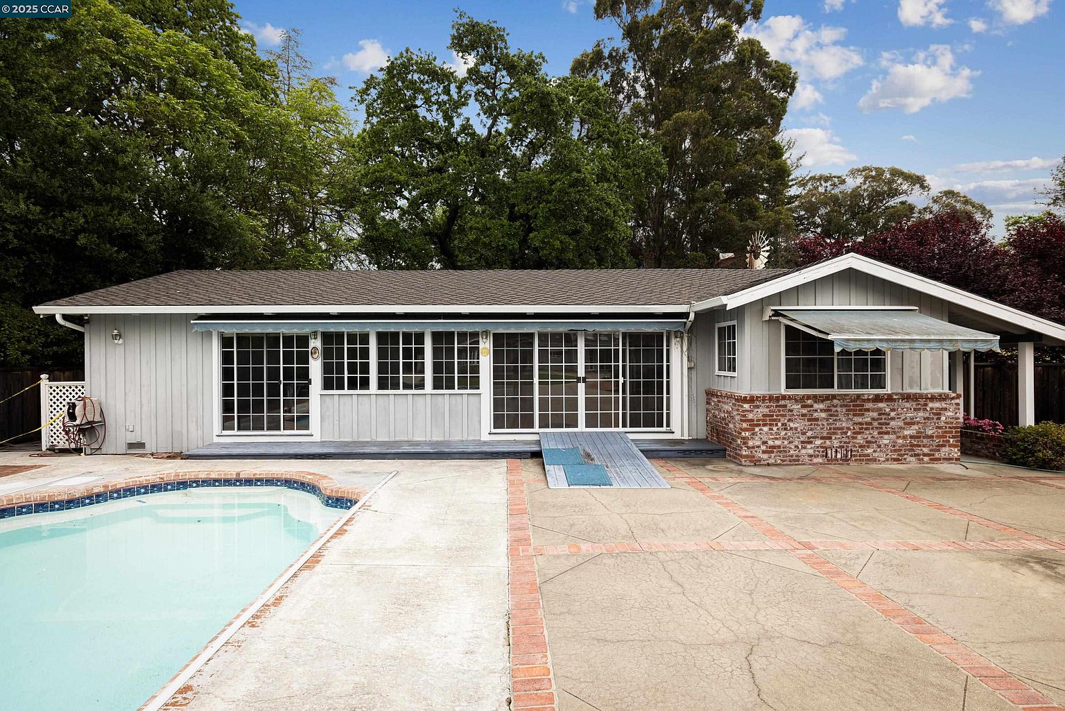 This is a rear view of a single-story house featuring a swimming pool and a patio area. The house has light gray siding, white-framed windows, and a dark gray roof. The patio is a mix of concrete and brick, providing a spacious outdoor living area adjacent to the pool.