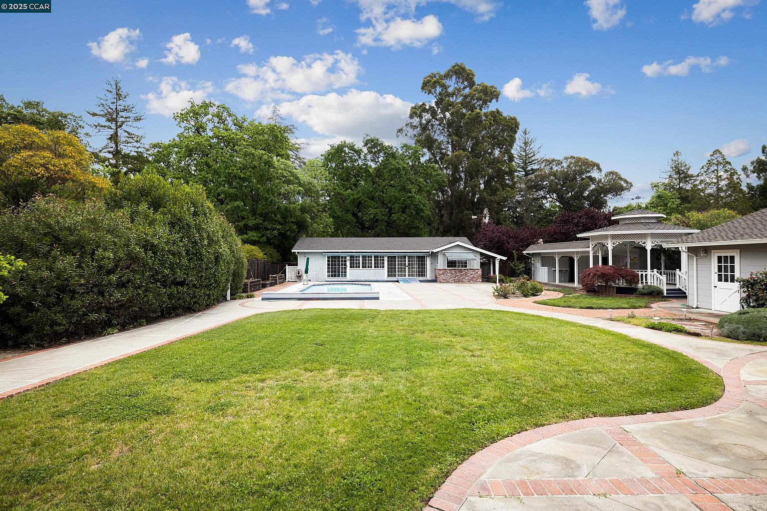 This exterior shot showcases a well-maintained yard with a lush green lawn, bordered by a brick-lined pathway. In the background, a single-story house with a pool and a gazebo-like structure are visible, surrounded by mature trees and landscaping. The image conveys a sense of tranquility and spaciousness, highlighting the property's outdoor living potential.