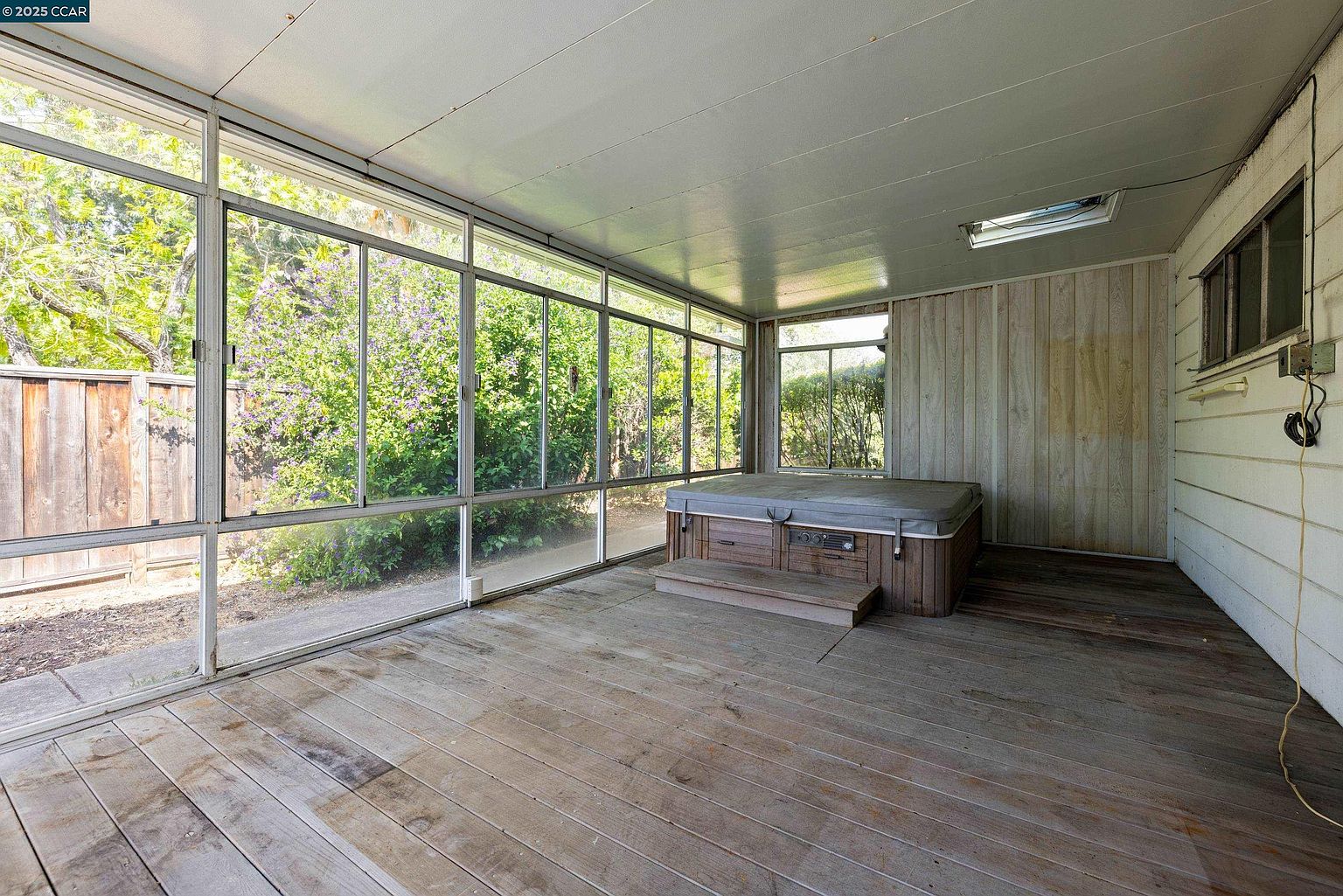 This sunroom features a hot tub, surrounded by large windows that offer views of the outdoor greenery. The room has a rustic feel with wooden floors and paneled walls, creating a relaxing and enclosed space. The natural light floods the room, enhancing the connection to the outdoors.
