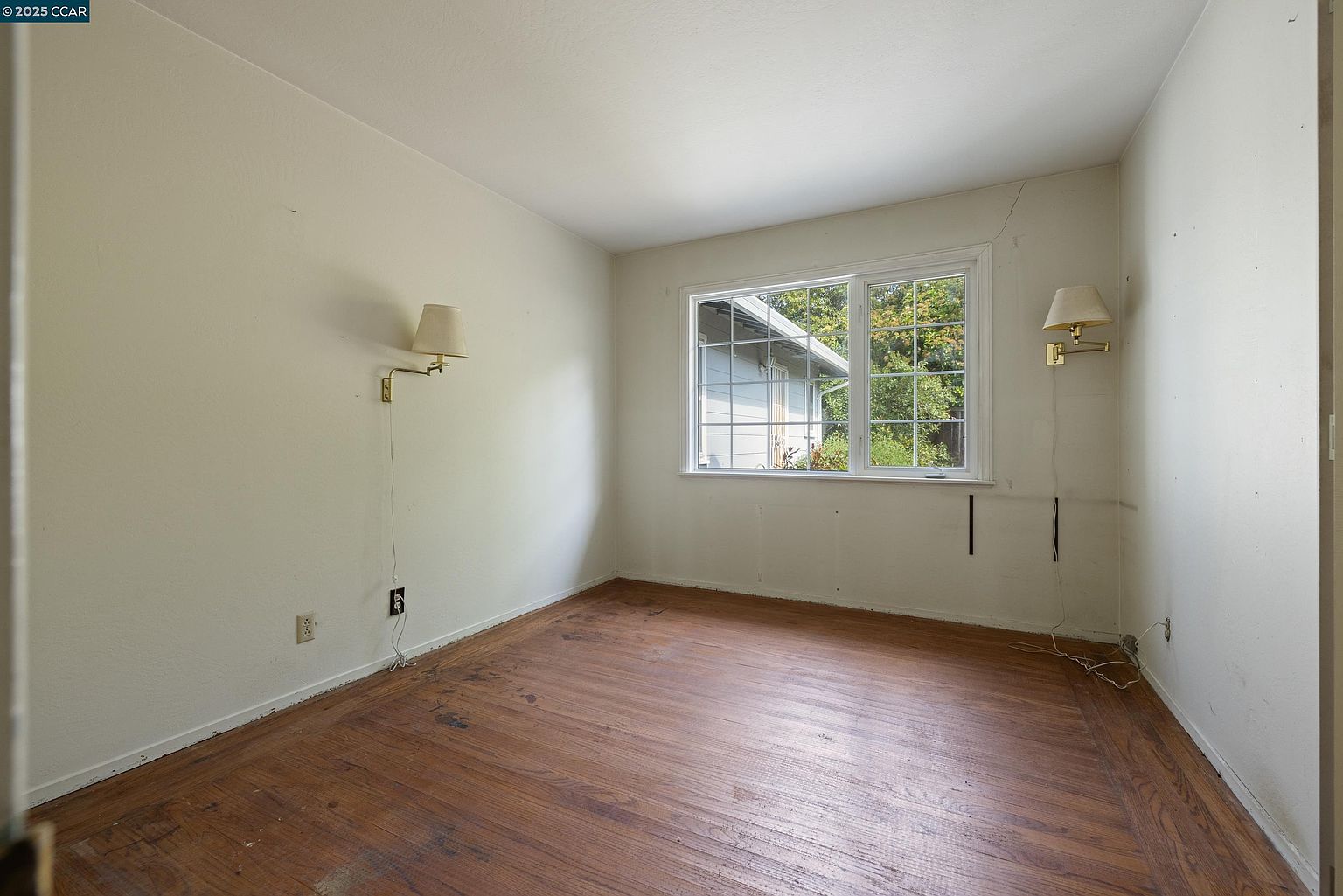 This is an interior shot of a bedroom featuring hardwood floors and light-colored walls. A window provides natural light, and there are wall-mounted lamps on either side. The room appears to be empty and in need of some updating.