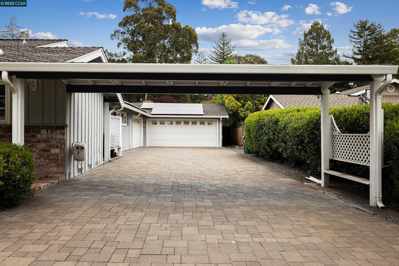 This image showcases a spacious driveway leading to a two-car garage, with a carport providing covered parking. The driveway is paved with interlocking bricks, adding texture and visual appeal. The garage features solar panels on the roof, highlighting energy efficiency, and is flanked by well-maintained landscaping, enhancing the property's curb appeal.