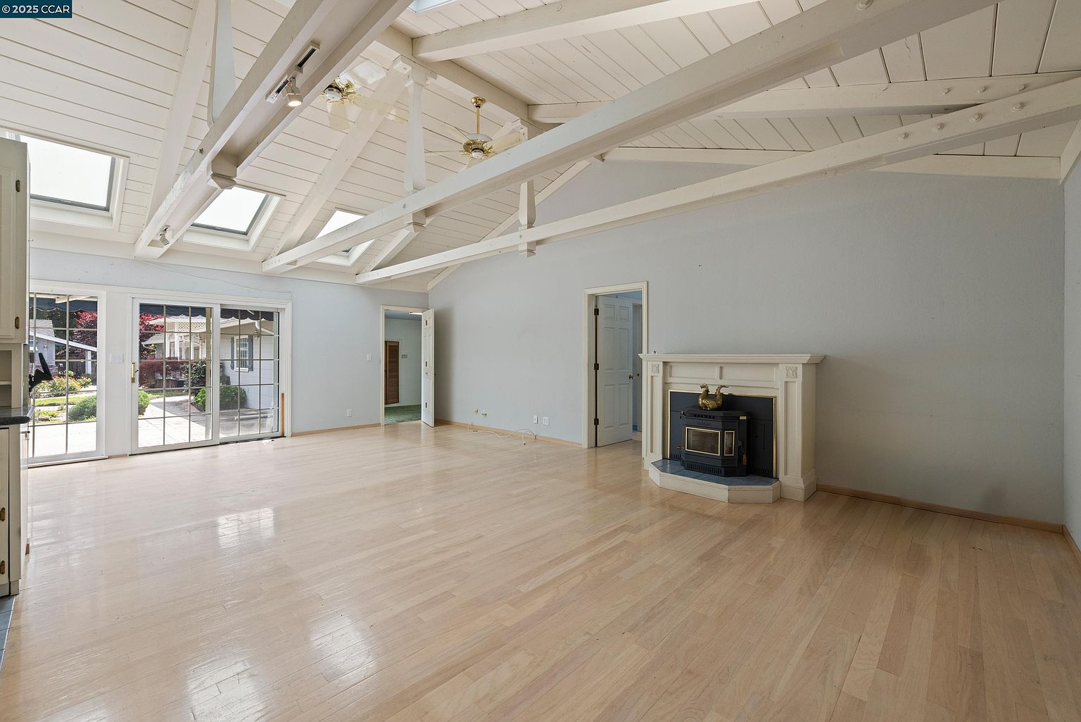 This is a spacious living room featuring light hardwood floors, a vaulted ceiling with exposed beams and skylights, and a fireplace with a white mantel. Sliding glass doors lead to the outside, providing ample natural light. The room is painted in a neutral color, creating a bright and airy atmosphere.