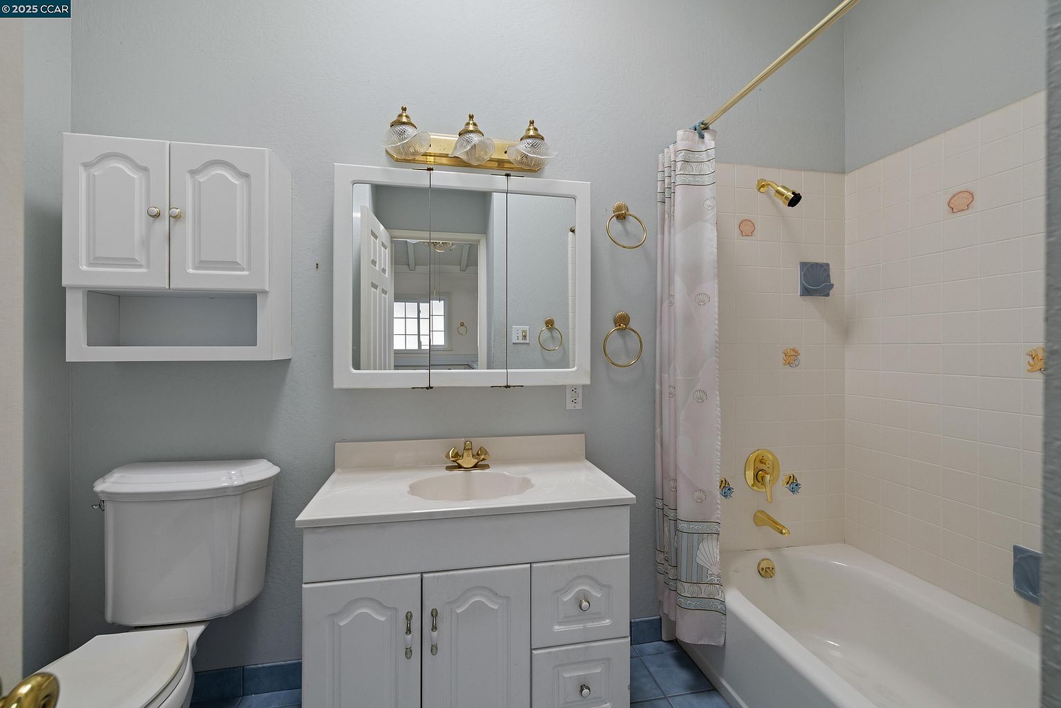 This is a bathroom featuring a white vanity with a sink, a mirror with lighting above, and a toilet. The walls are painted a light blue, and the floor is tiled in blue. The bathtub/shower combination has a shower curtain and decorative seashell accents on the tiled wall, creating a vintage, beach-themed aesthetic.