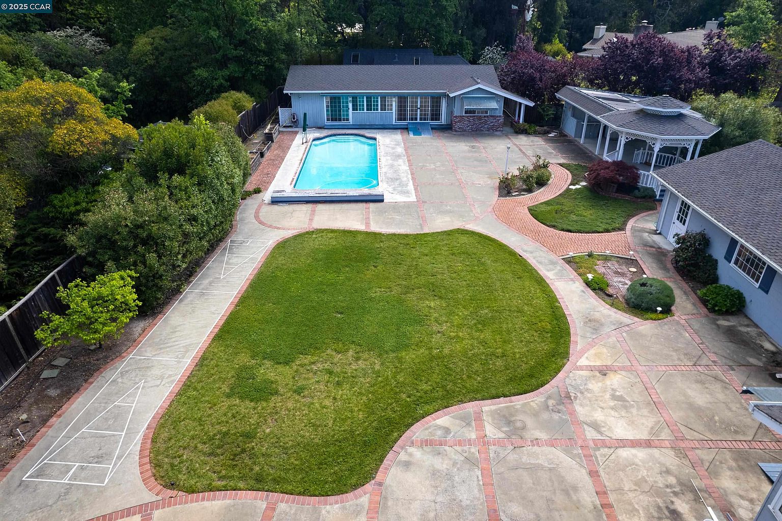 This aerial view showcases a well-maintained property featuring a light blue single-story home with a dark roof, a swimming pool, a gazebo, and a spacious green lawn. The property is surrounded by mature trees and landscaping, creating a private and serene atmosphere. The layout includes paved areas with brick accents, adding to the overall aesthetic appeal.