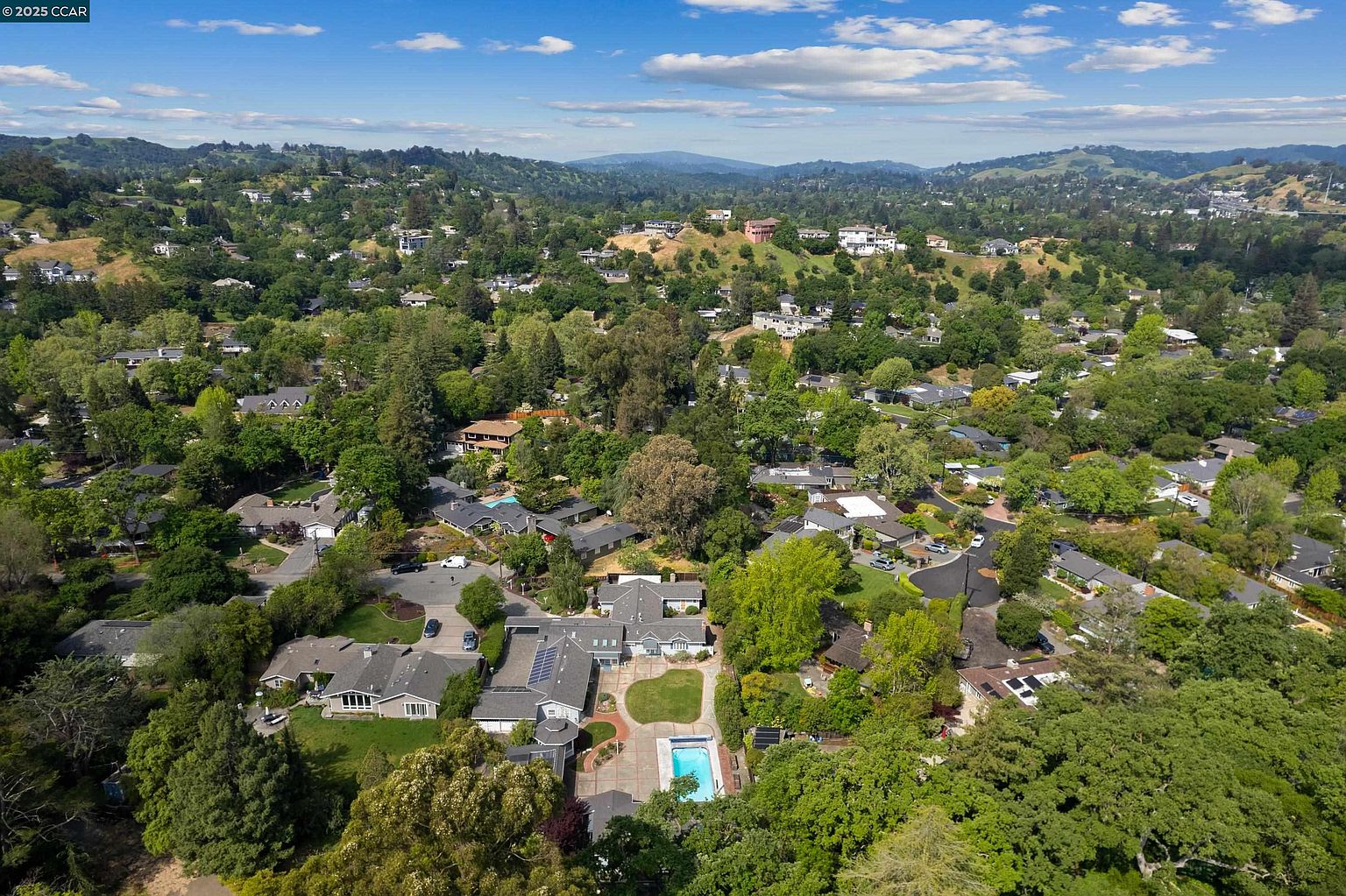 This aerial view showcases a neighborhood nestled among lush green hills and trees. Several houses with varying architectural styles are visible, some featuring pools and well-maintained yards. The overall impression is one of a peaceful and affluent residential area.