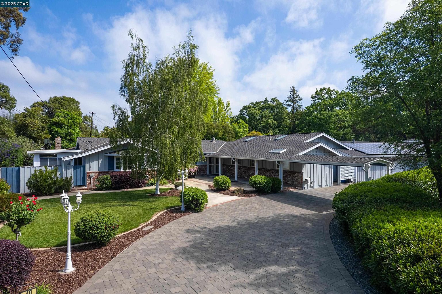 This is a front view of a single-story home with a well-maintained yard and a brick driveway. The house features a combination of light blue siding and brick accents, with a gray roof. Mature trees and landscaping add to the property's curb appeal, creating a welcoming and established feel.