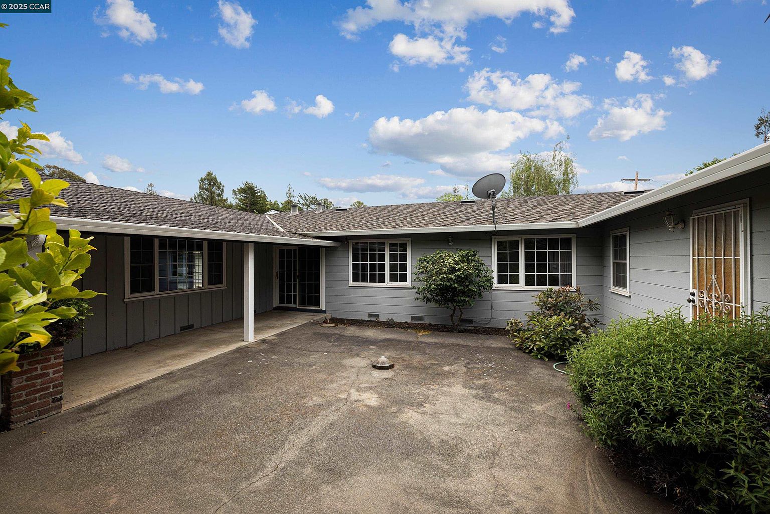 This is a rear view of a single-story house with a gray exterior and a dark gray roof. The house features multiple windows with white frames and a paved courtyard area. There are various plants and shrubs around the courtyard, adding a touch of greenery to the scene.